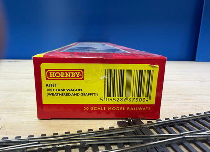 A red Hornby model railway box for a 100t tank wagon, shown on a light wood surface with model train tracks in the foreground and a blue background.